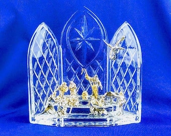 May include: A clear glass nativity scene with a diamond pattern and gold and silver figurines. The scene features a star, angels, and figures of the nativity story. The background is a solid blue.