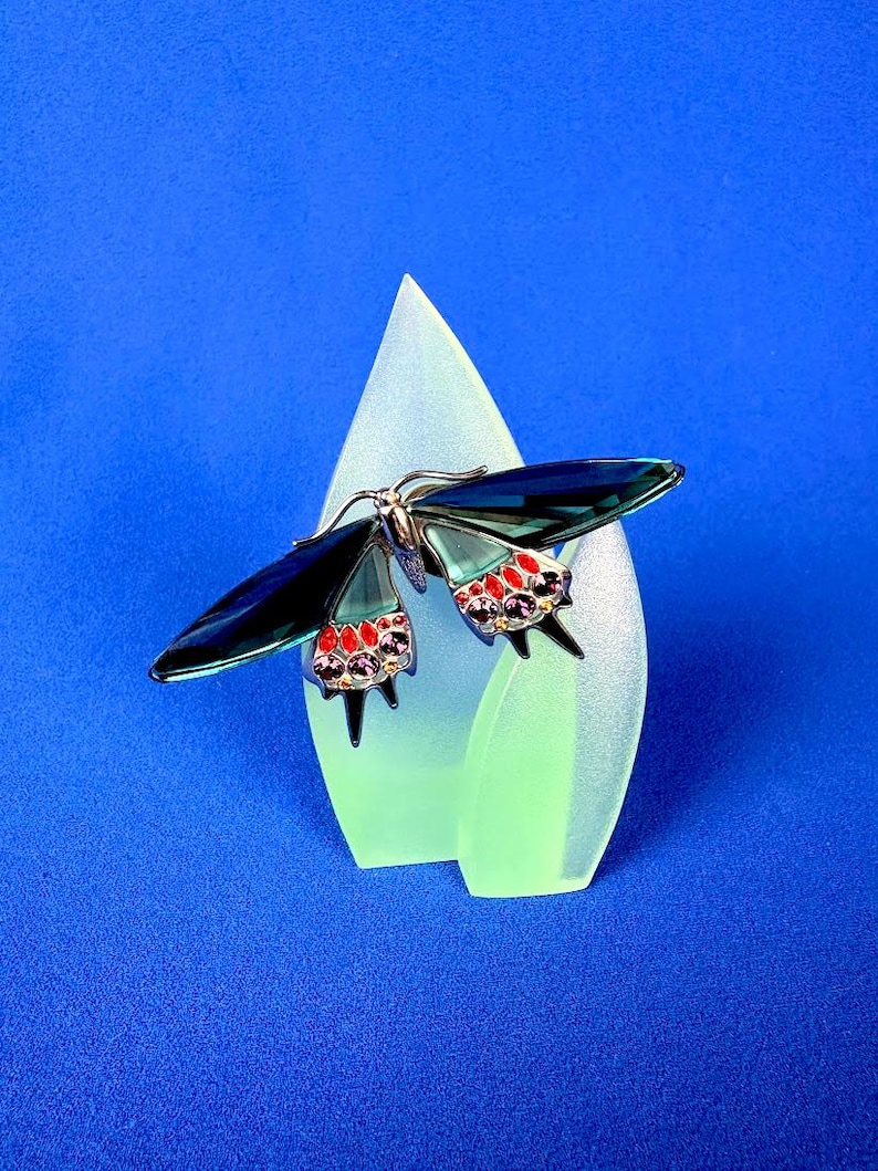 Puede incluir: Broche decorativo de mariposa con alas azul oscuro y detalles rojos y morados. El broche est&aacute; colocado sobre una pantalla en forma de l&aacute;grima verde claro sobre un fondo azul. El cuerpo de la mariposa es plateado.