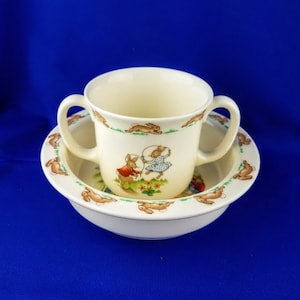 May include: A white ceramic children's bowl and cup set with a bunny rabbit design. The bowl and cup have two handles. The design features bunnies playing with a hoop.