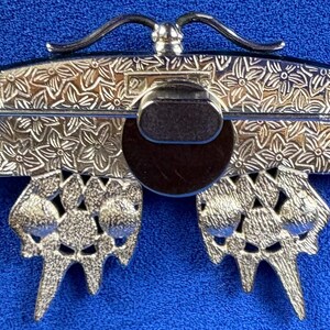 Puede incluir: Broche de mariposa plateada con intrincados detalles florales en las alas y el cuerpo. El broche tiene un cierre de alfiler en la parte posterior y un centro marr&oacute;n oscuro. Las alas de la mariposa tienen un dise&ntilde;o calado texturizado.