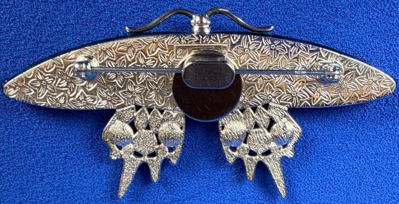 Puede incluir: Broche de mariposa plateada con intrincados detalles florales en las alas y el cuerpo. El broche tiene un cierre de alfiler en la parte posterior y un centro marr&oacute;n oscuro. Las alas de la mariposa tienen un dise&ntilde;o calado texturizado.