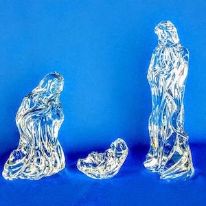 May include: A clear glass nativity scene set against a blue background. The set includes three figures: a kneeling figure, a standing figure, and a baby in a manger. The figures have a textured, flowing design.