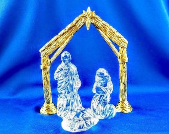 May include: A clear glass nativity scene with gold-colored accents. The scene includes figures of Mary, Joseph, and the baby Jesus under a gold-colored wooden-style structure. The background is a solid blue.