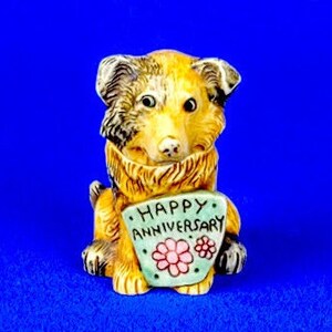 May include: A ceramic figurine of a brown and black dog holding a green card with the text "Happy Anniversary" and two pink flowers.