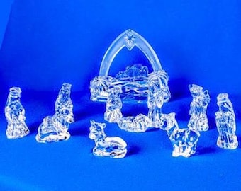 May include: A clear glass nativity scene set against a blue background. The set includes figures of the holy family, animals, and the three wise men. The figures are detailed and reflect light.