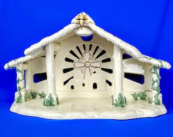 May include: A white ceramic nativity scene with a star on top and a cross in the center. The scene is decorated with green ivy and has a white background.