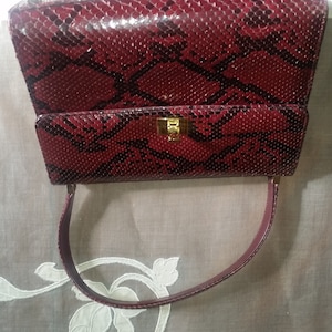 May include: A burgundy snakeskin handbag with a gold clasp and a single top handle.