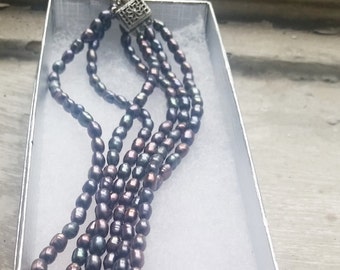 Può includere: Una collana multifilo realizzata con perle scure e iridescenti. La collana è in una scatola bianca su uno sfondo bianco.