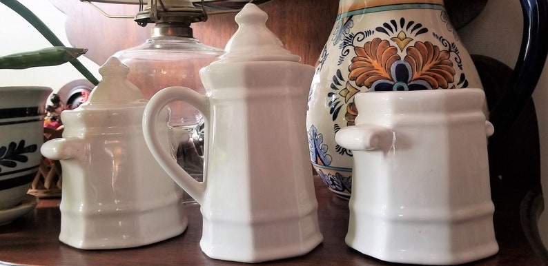 Puede incluir: Tres recipientes de cer&aacute;mica blanca con tapas, dos con asas y uno con tapa. Los recipientes est&aacute;n sobre una superficie de madera junto a una jarra de cer&aacute;mica azul y blanca con dise&ntilde;os florales.