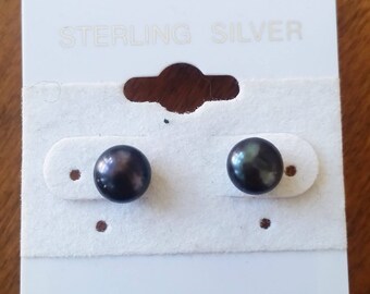 Puede incluir: Un par de pendientes de perlas grises oscuras en una tarjeta blanca. La tarjeta dice "STERLING SILVER" en letras plateadas. Los pendientes son redondos y tienen un brillo iridiscente sutil.