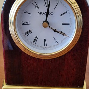 Peut inclure: Une horloge à quartz en bois avec une lunette dorée et des chiffres romains. Le cadran est blanc avec le nom de la marque "MOVADO" imprimé. Le cadre est en bois marron foncé et la base dorée.