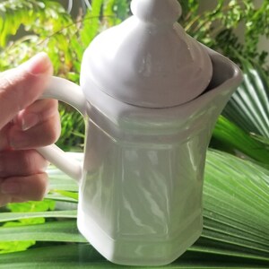Puede incluir: Una jarra de crema blanca de cer&aacute;mica con tapa y asa. La jarra tiene un dise&ntilde;o texturizado y est&aacute; sobre una hoja verde.