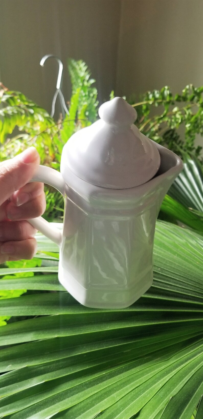 Puede incluir: Una jarra de crema blanca de cer&aacute;mica con tapa y asa. La jarra tiene un dise&ntilde;o texturizado y est&aacute; sobre una hoja verde.