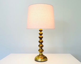 Könnte beinhalten: Eine goldene Tischlampe mit einem weißen Stofflampenschirm. Der Lampenfuß hat eine Reihe von runden, dekorativen Elementen.