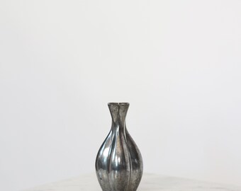 Könnte beinhalten: Eine silberfarbene Vase mit bauchigem Körper und schmalem Hals. Die Vase hat vertikale Rillen und einen metallischen Glanz, der das Licht reflektiert. Die Vase ist ein dekorativer Gegenstand.