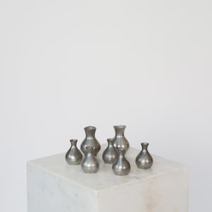 May include: A collection of seven small, silver-colored vases with rounded bodies and narrow necks. The vases are arranged on a white marble cube, set against a plain white background. The vases vary slightly in size and shape.