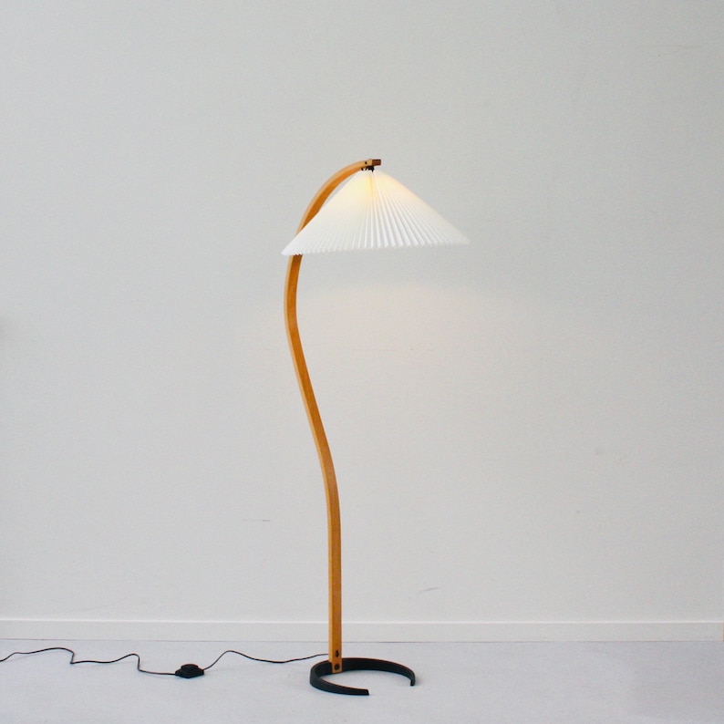 K&ouml;nnte beinhalten: Eine Stehlampe aus Holz mit einem wei&szlig;en, gefalteten Lampenschirm. Die Lampe hat einen gebogenen Holzfu&szlig; und einen schwarzen Fu&szlig;.