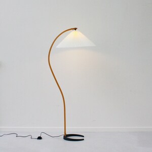 K&ouml;nnte beinhalten: Eine wei&szlig;e Stehlampe mit einem gebogenen Holzarm und einem wei&szlig;en, gefalteten Lampenschirm. Die Lampe hat einen schwarzen Sockel.
