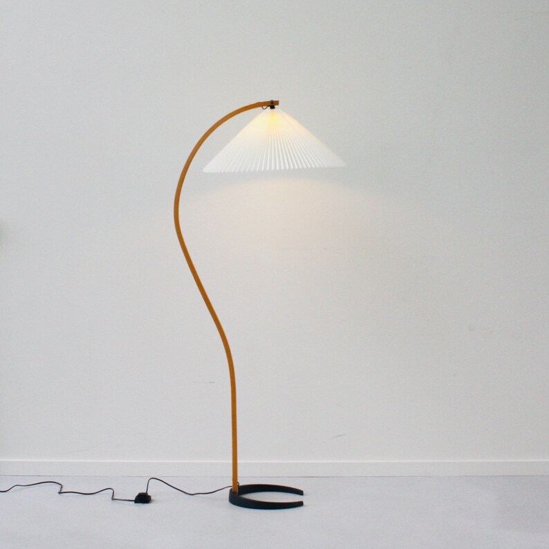 K&ouml;nnte beinhalten: Eine wei&szlig;e Stehlampe mit einem gebogenen Holzarm und einem wei&szlig;en, gefalteten Lampenschirm. Die Lampe hat einen schwarzen Sockel.