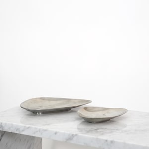 May include: Two silver-toned, organically shaped serving dishes. The larger dish is elongated, while the smaller one is more rounded. Both are placed on a white marble surface, creating a minimalist aesthetic. The dishes appear to be made of metal.
