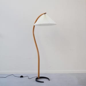K&ouml;nnte beinhalten: Eine Stehlampe aus Holz mit einem gebogenen Holzarm und einem wei&szlig;en, gefalteten Lampenschirm. Die Lampe hat ein schwarzes Kabel und einen schwarzen Sockel.