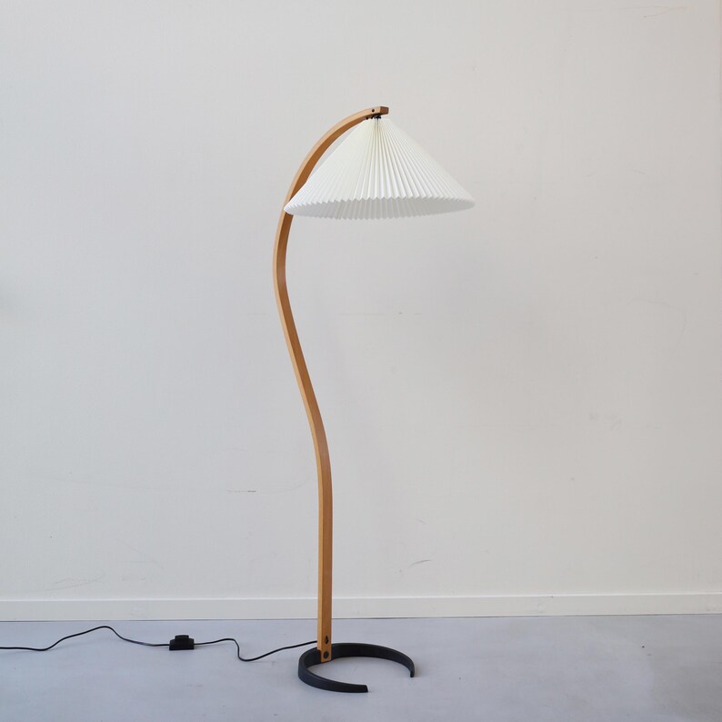 K&ouml;nnte beinhalten: Eine Stehlampe aus Holz mit einem gebogenen Holzarm und einem wei&szlig;en, gefalteten Lampenschirm. Die Lampe hat ein schwarzes Kabel und einen schwarzen Sockel.