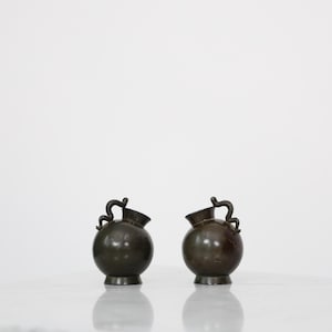 May include: Two antique bronze-coloured vases with a round body and a small spout. Each vase has a decorative handle. The vases are displayed on a white surface.