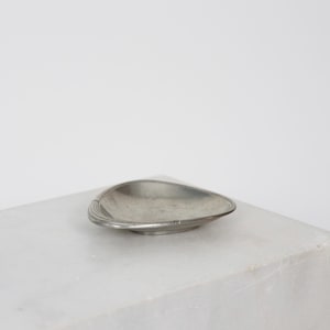 May include: A small, antique, silver-colored dish with a unique, asymmetrical shape. The dish has a slightly raised rim and decorative lines along one side. The dish is resting on a white surface.