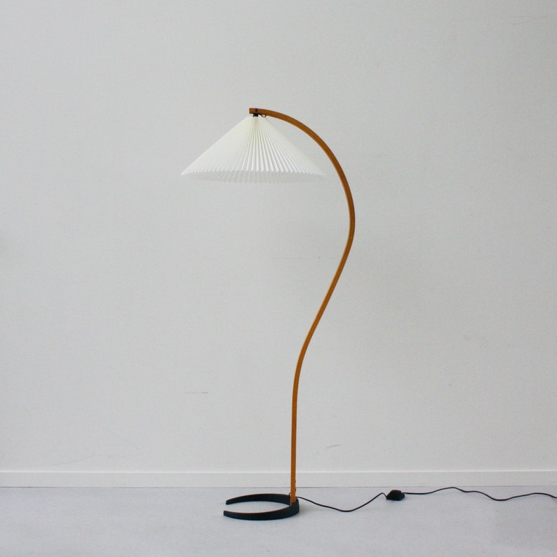 K&ouml;nnte beinhalten: Eine gebogene Stehlampe aus Holz mit einem wei&szlig;en, gefalteten Lampenschirm und einem schwarzen Fu&szlig;.