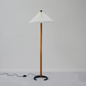 K&ouml;nnte beinhalten: Eine wei&szlig;e, gefaltete Lampenleuchte mit einem Holzstiel und einem schwarzen Metallfu&szlig;. Die Lampe steht auf einem wei&szlig;en Boden.