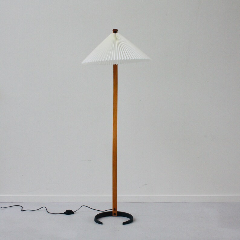 K&ouml;nnte beinhalten: Eine wei&szlig;e, gefaltete Lampenleuchte mit einem Holzstiel und einem schwarzen Metallfu&szlig;. Die Lampe steht auf einem wei&szlig;en Boden.