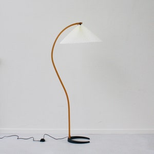 K&ouml;nnte beinhalten: Eine gebogene Holzstehlampe mit einem wei&szlig;en, gefalteten Stoffschirm. Die Lampe hat einen schwarzen Sockel und ein Kabel mit einem Fu&szlig;schalter.