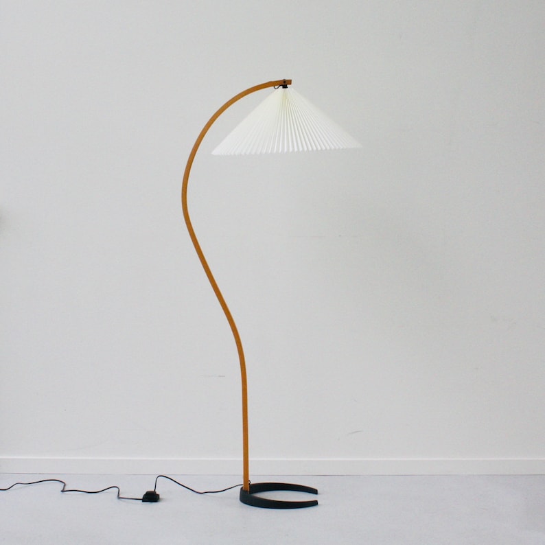 K&ouml;nnte beinhalten: Eine gebogene Holzstehlampe mit einem wei&szlig;en, gefalteten Stoffschirm. Die Lampe hat einen schwarzen Sockel und ein Kabel mit einem Fu&szlig;schalter.