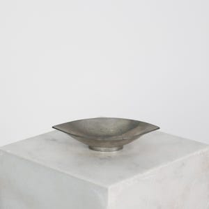 May include: A small, gray metal bowl with a unique, angular shape. The bowl has a shallow depth and sits on a circular base. The bowl is displayed on a white, marble-like surface against a plain white background.