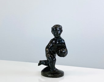 Könnte beinhalten: Eine schwarze Keramikfigur eines Cherubs, der eine Kugel hält. Der Cherub steht auf einem runden Sockel.