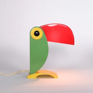 May include: A green and yellow toucan-shaped table lamp with a red shade. The lamp is switched on and the light is shining.