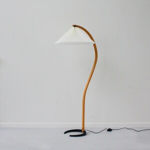 K&ouml;nnte beinhalten: Eine gebogene Stehlampe aus Holz mit einem wei&szlig;en, gefalteten Lampenschirm und einem schwarzen Fu&szlig;.