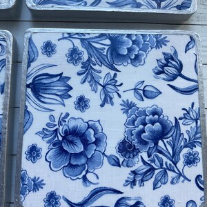May include: Four square blue and white ceramic coasters with a floral pattern. The coasters have a silver trim around the edges.