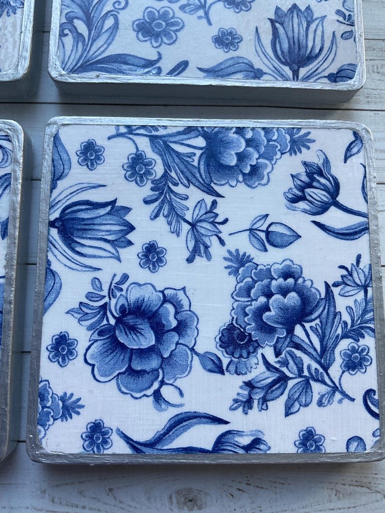 May include: Four square blue and white ceramic coasters with a floral pattern. The coasters have a silver trim around the edges.