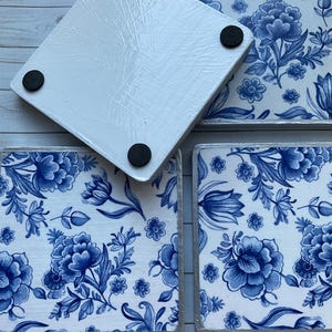 May include: Four square coasters with a blue and white floral pattern. The coasters have a white background with blue flowers and leaves. The coasters have a silver trim around the edges. The bottom of the coasters have black rubber feet.
