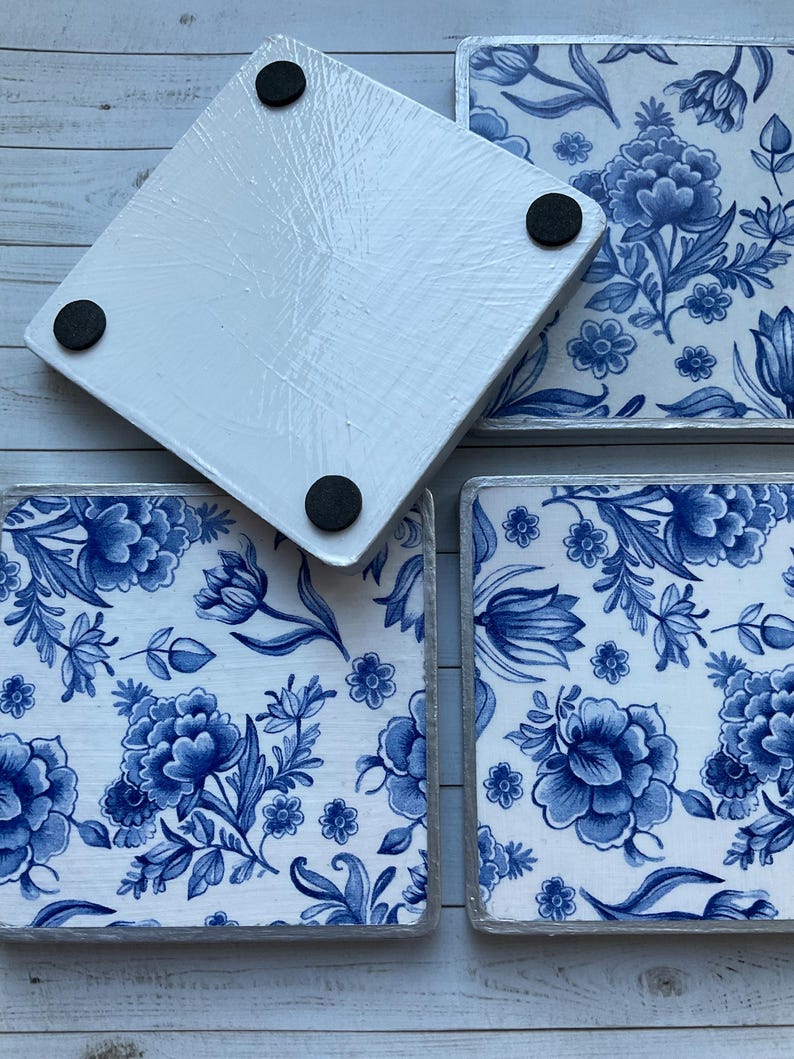 May include: Four square coasters with a blue and white floral pattern. The coasters have a white background with blue flowers and leaves. The coasters have a silver trim around the edges. The bottom of the coasters have black rubber feet.
