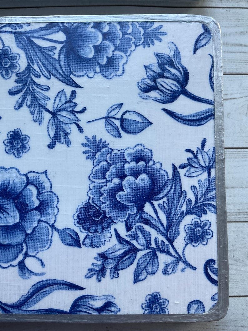 May include: A square ceramic tile with a blue and white floral pattern. The tile has a silver border.