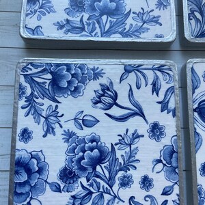 May include: Four square blue and white ceramic coasters with a floral pattern. The coasters have a silver trim around the edges.