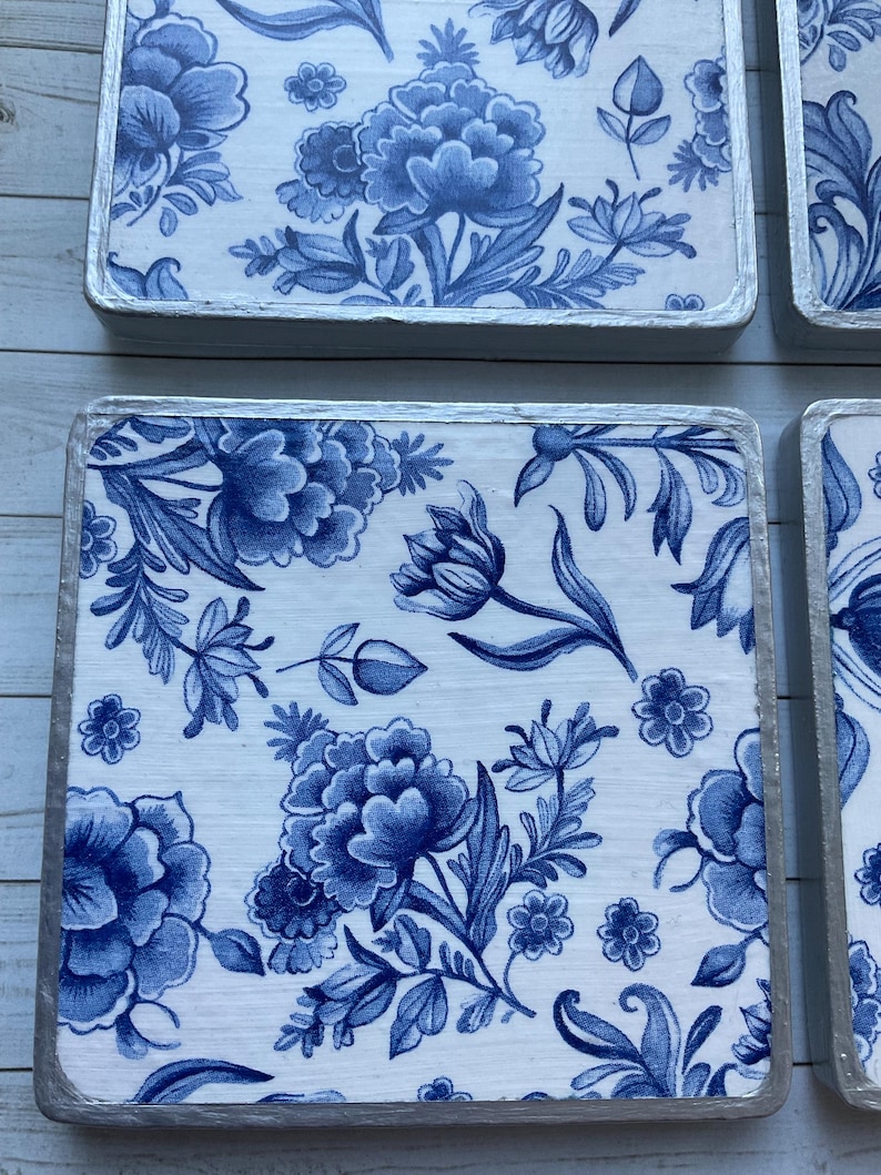 May include: Four square blue and white ceramic coasters with a floral pattern. The coasters have a silver trim around the edges.