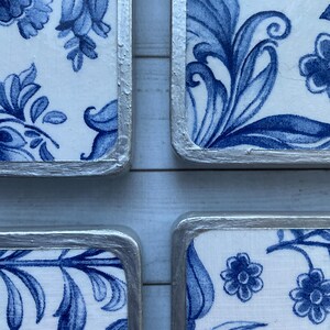 May include: Four square ceramic coasters with a blue and white floral pattern. The coasters have a silver trim around the edges.