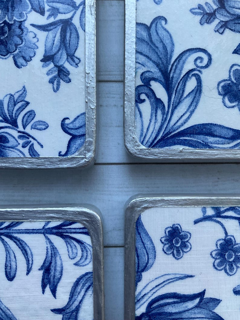 May include: Four square ceramic coasters with a blue and white floral pattern. The coasters have a silver trim around the edges.