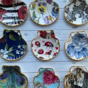 May include: Nine decorative seashells with gold trim, each featuring a different patterned design. The designs include floral patterns, ladybugs, peacocks, dogs, and a rose.