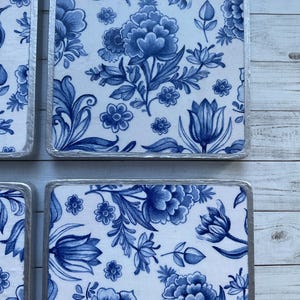 May include: Four square blue and white ceramic coasters with a floral pattern. The coasters have a silver trim.