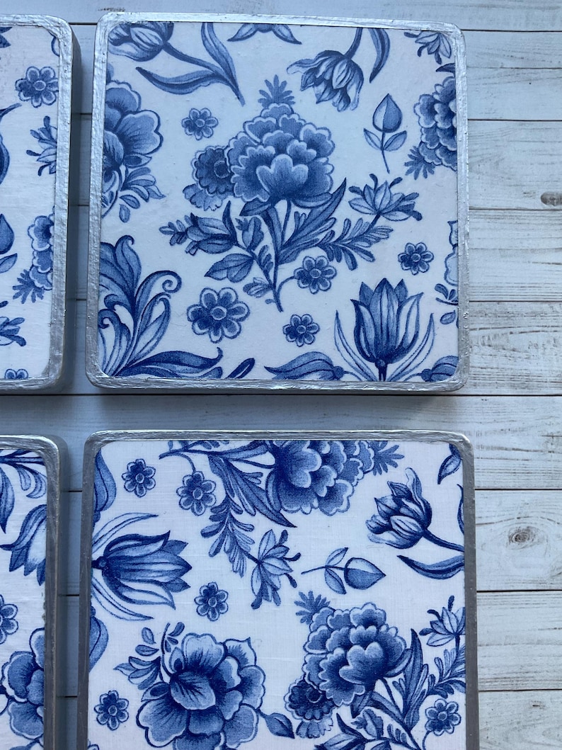 May include: Four square blue and white ceramic coasters with a floral pattern. The coasters have a silver trim.