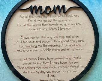 May include: A circular wooden plaque with a black frame, featuring the word "mom" in a cursive script. The plaque displays a poem dedicated to a mother, with the names "Amanda & Sam" at the base.